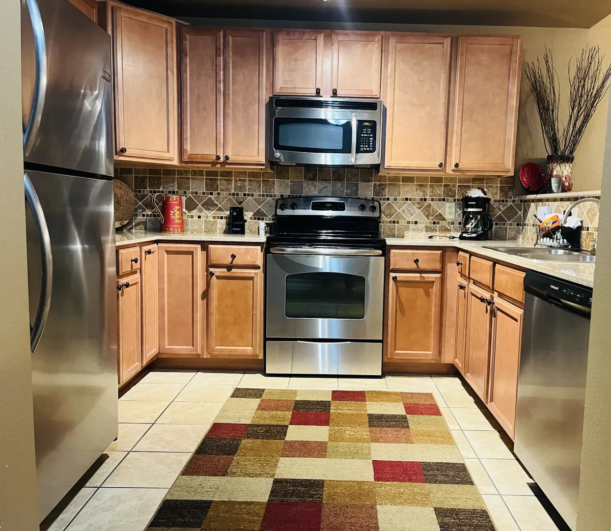 A modern kitchen with stainless steel appliances, polished granite countertops, a colorful rug, and decorative wall accents. The kitchen exudes warmth and functionality, with plenty of workspace ideal for preparing anything from family feasts to quick snacks.