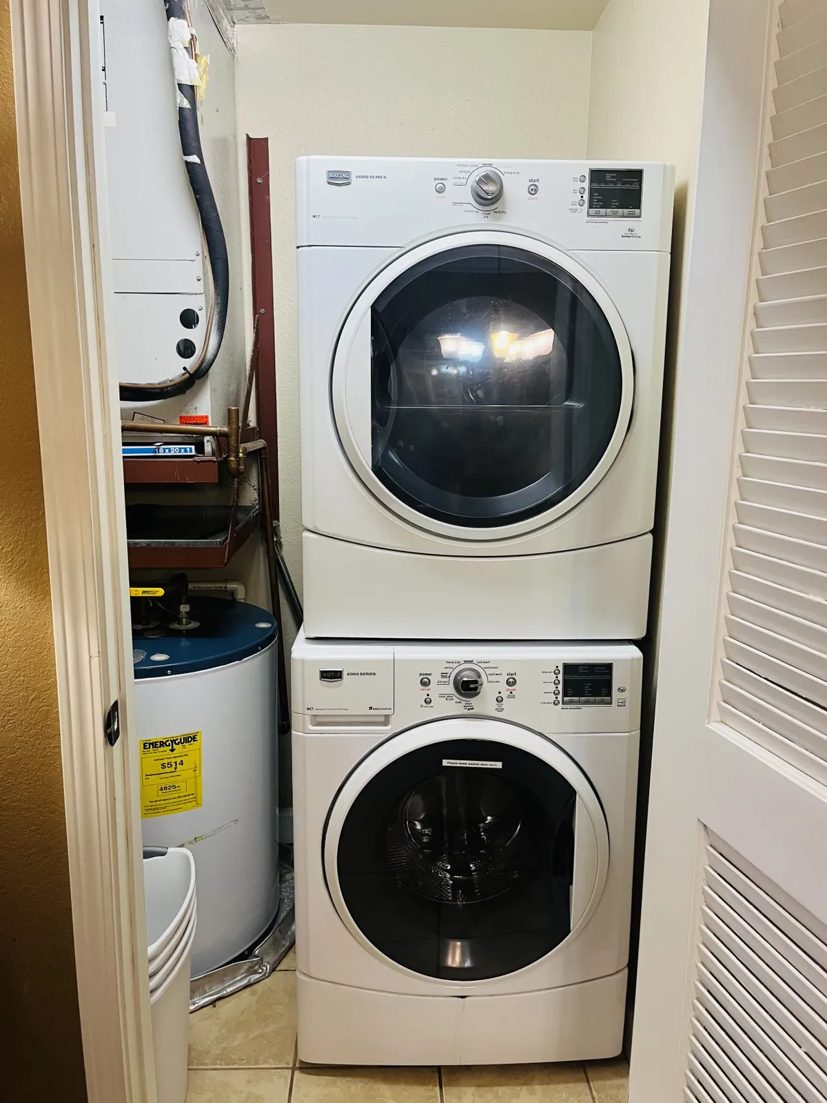A compact yet highly practical laundry area featuring a stacked washer and dryer, water heater, and storage bins. The layout is designed for easy organization and quick access to laundry essentials.