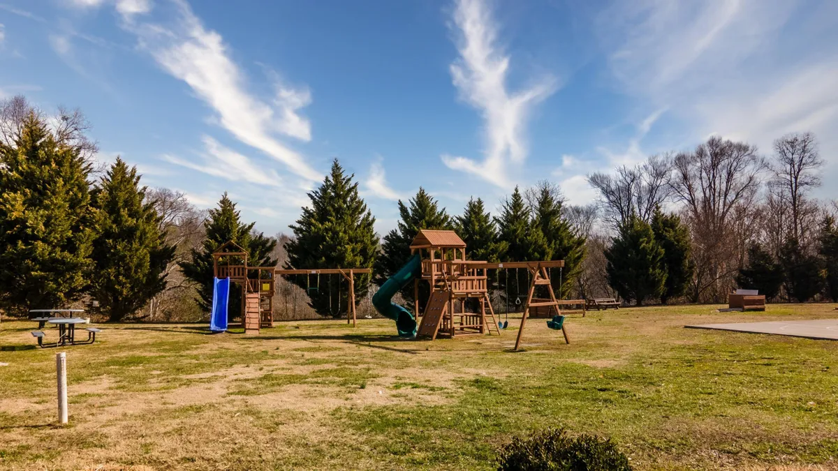 A colorful playground equipped with swings, a slide, and a nearby picnic area. Children are playing while families gather for picnics and casual socializing. The area is lively and safe, surrounded by greenery and open space.