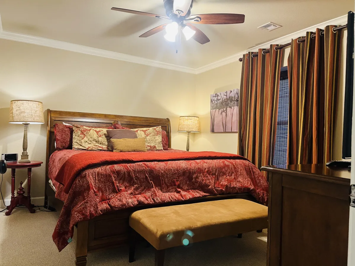A bedroom with a bed dressed in red bedding, a nightstand with a lamp, striped curtains letting in natural light, and an art piece on the wall creates a warm and inviting atmosphere. The layout suggests comfort and relaxation, with thoughtful decor and soft lighting.