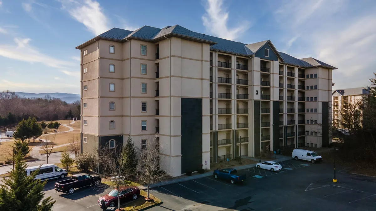 A wide view of the multi-story condo building surrounded by lush greenery and ample parking. The balconies are visible, offering a glimpse of the elevated outdoor spaces, while the setting sun casts a warm glow, creating a welcoming atmosphere.
