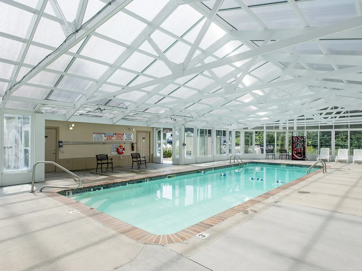 This image showcases a spacious indoor swimming pool area with white wooden beams overhead and several lounge chairs lining the poolside. The atmosphere is bright and inviting, emphasizing leisure and relaxation in a sheltered environment, regardless of the weather outside.