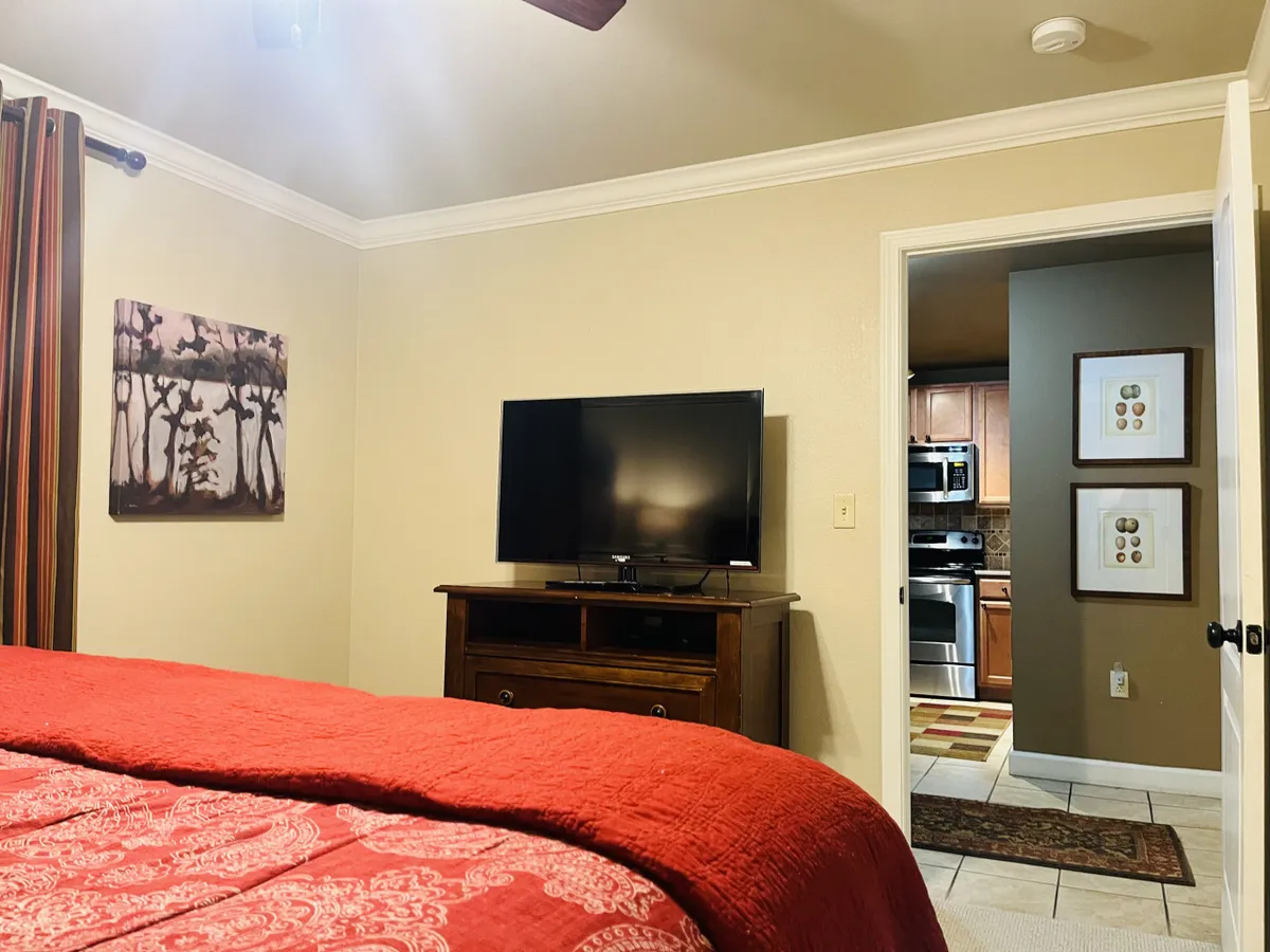 A comfortable master suite with red quilted bedding, a flat-screen TV, art on the walls, and a view into the adjacent kitchen. The space is cozy and welcoming, perfect for relaxing after a day of adventure or an evening at a local show.