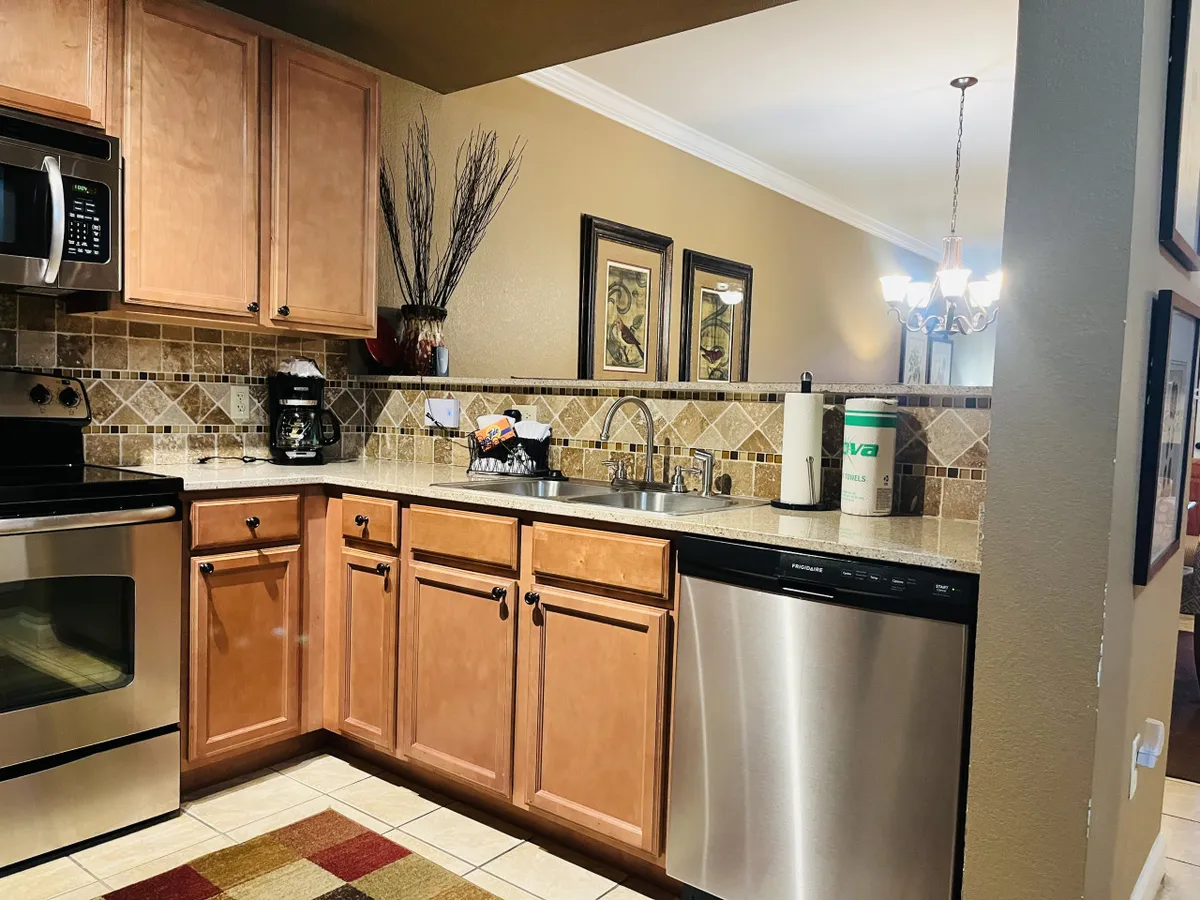 A bright kitchen space featuring stainless steel appliances, wooden cabinets, decorative wall art, and a spacious countertop. The room looks inviting and functional, with modern amenities clearly visible. The arrangement suggests ample room for both cooking and entertaining guests.
