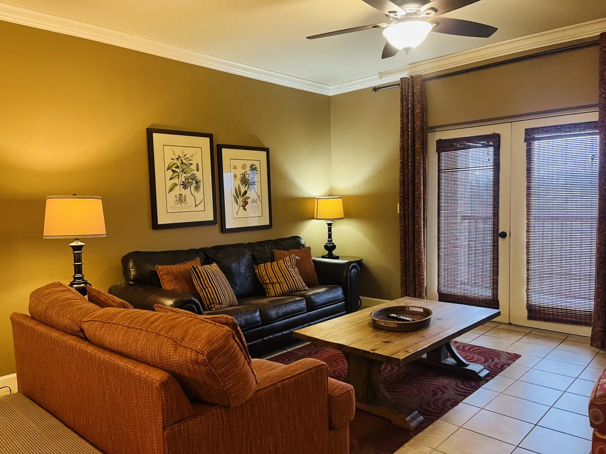 A cozy living room with a brown leather sofa, orange armchair, wooden coffee table, and framed botanical prints. The arrangement suggests a social, comfortable space, and the layout implies proximity to a balcony or window, hinting at scenic views beyond.