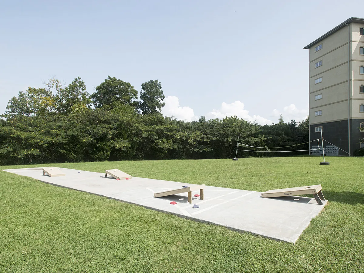 The image presents a lush, green recreation area equipped with bean bag toss boards and a volleyball net. The scene suggests group activities, play, and socializing, set in a safe and inviting open space ideal for families and children.